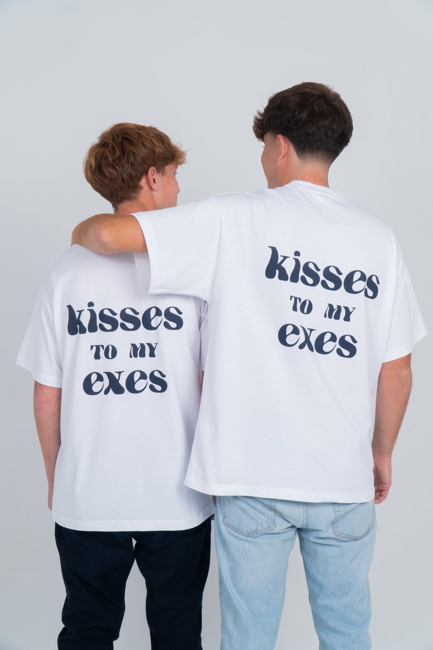 Dos chicos de espaldas, uno de ellos rodea con su brazo al otro por el hombro. Llevan la camiseta blanca con el diseño en la espalda de "kisses to my exes". Y en el pecho está el logo bordado de exes en azul ocuro. 