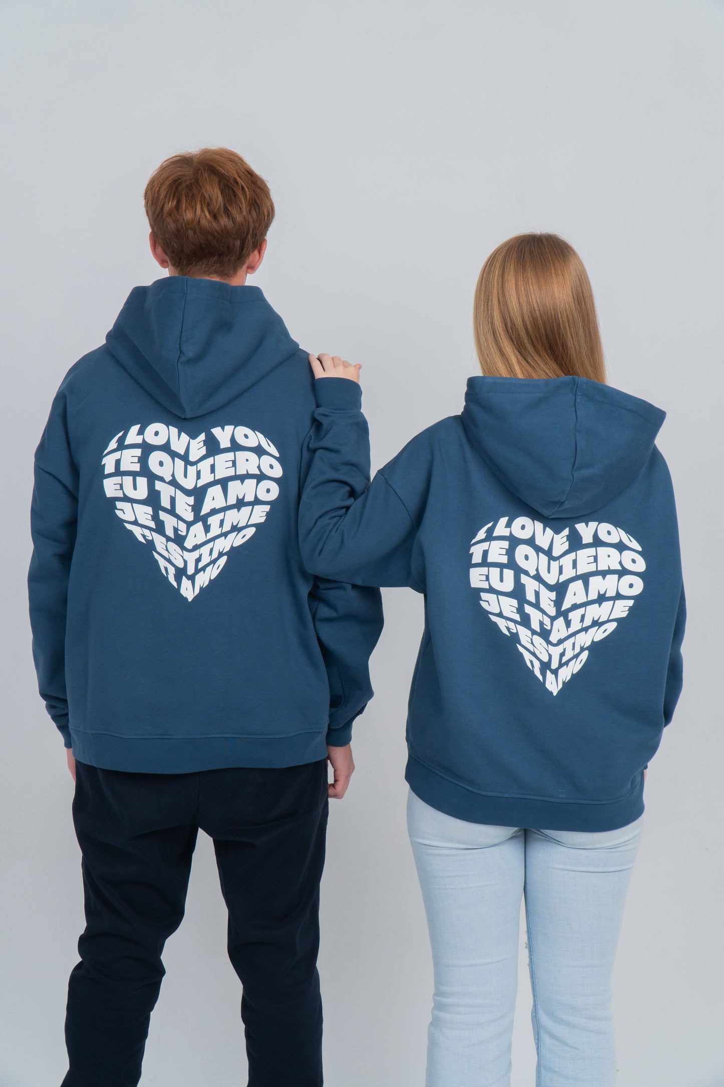 Chico y chica de espaldas. Chica tiene la mano encima del hombro de él. En la espalda de la sudadera azul se muestra el diseño de un corazón formado por la palabra te quiero en diferentes idiomas. El orden es: i love you, te quiero, eu te amo, je t'aime, t'estimo, ti amo. 