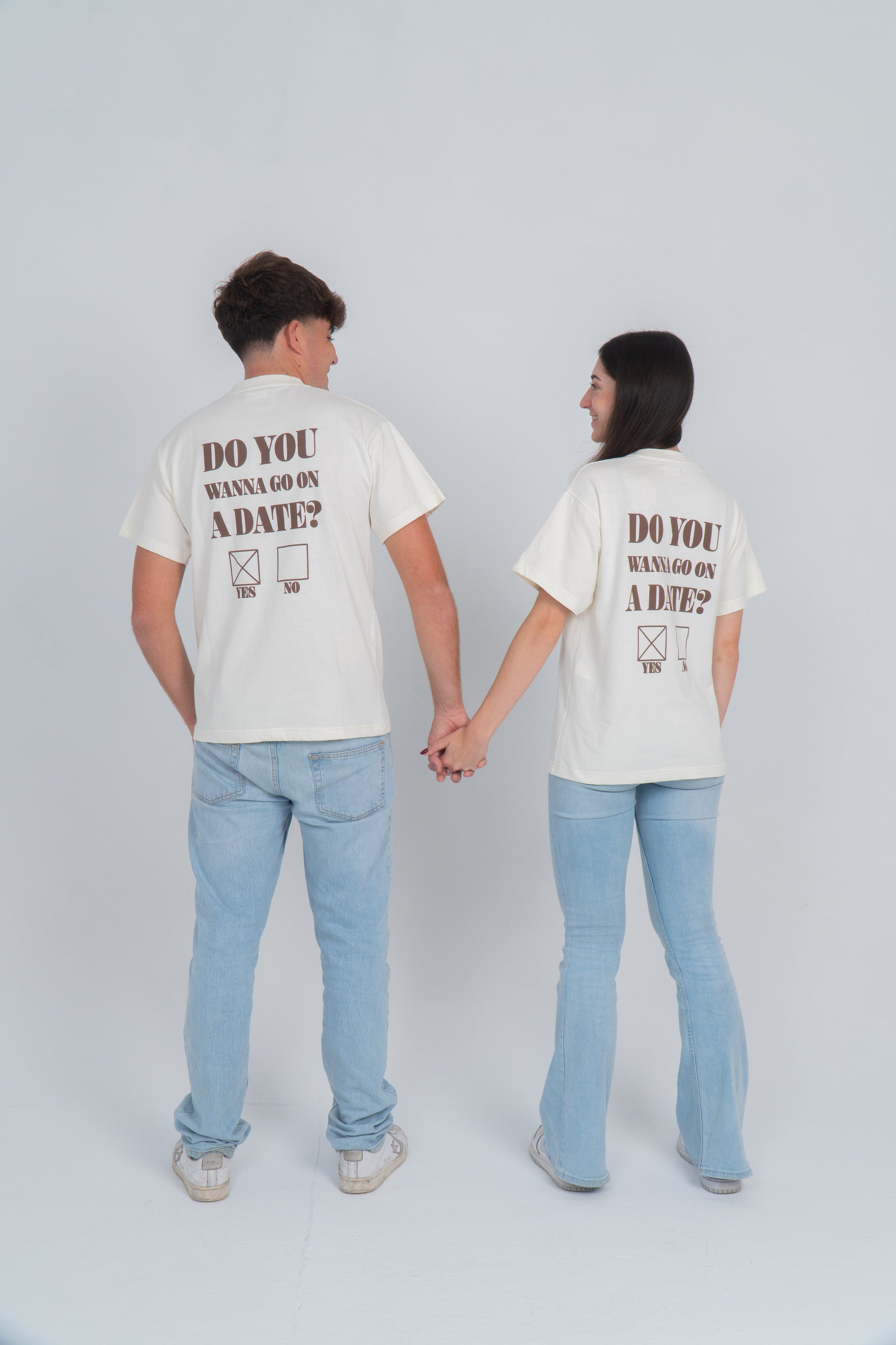 Chico y chica cogidos de la mano llevan la camiseta beige con el diseño en la espalda de do you wanna go on a date?