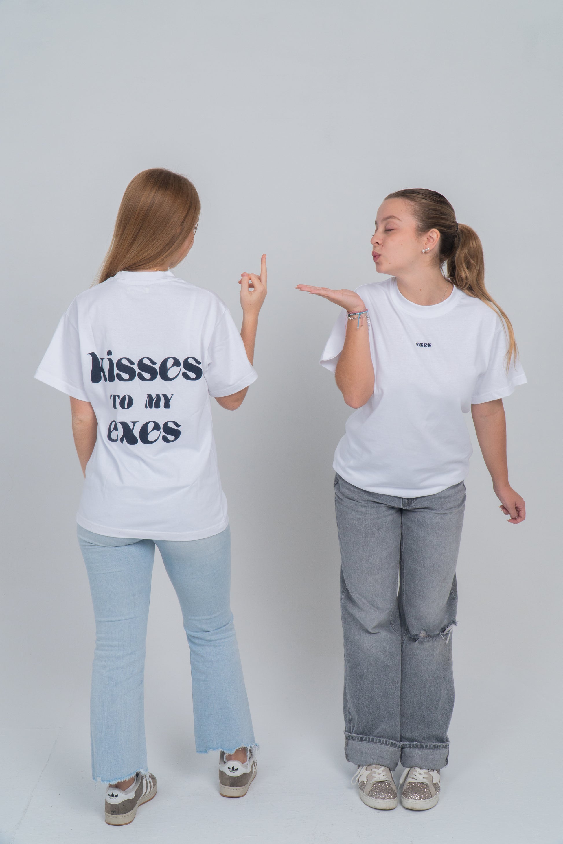 Dos chicas. Una de espalda y otra de cara. Una de ellas le saca el dedo del medio y la otra le lanza un beso. Llevan la camiseta blanca con el diseño en la espalda de "kisses to my exes". Y en el pecho está el logo bordado de exes en azul ocuro. 