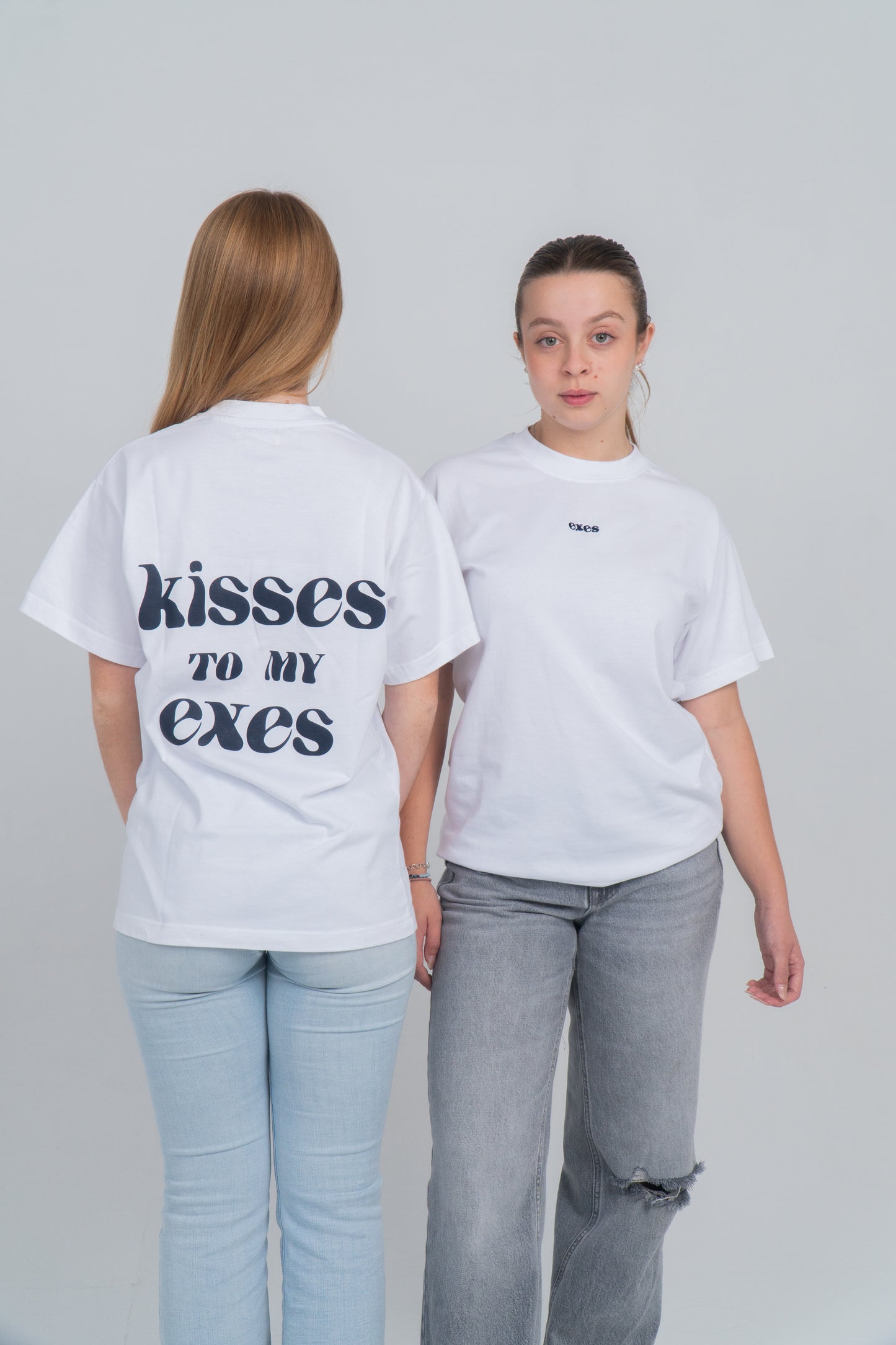 Dos chicas. Una de espalda y otra de cara. Llevan la camiseta blanca con el diseño en la espalda de "kisses to my exes". Y en el pecho está el logo bordado de exes en azul ocuro. 