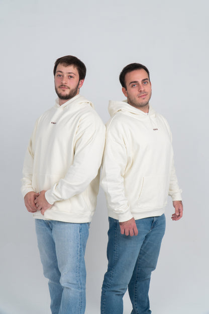 Dos chicos, están hombro con hombro, de cara. Llevan la hoodie/sudadera beige vemos el logo de la marca bordado en marrón oscuro en el centro del pecho.