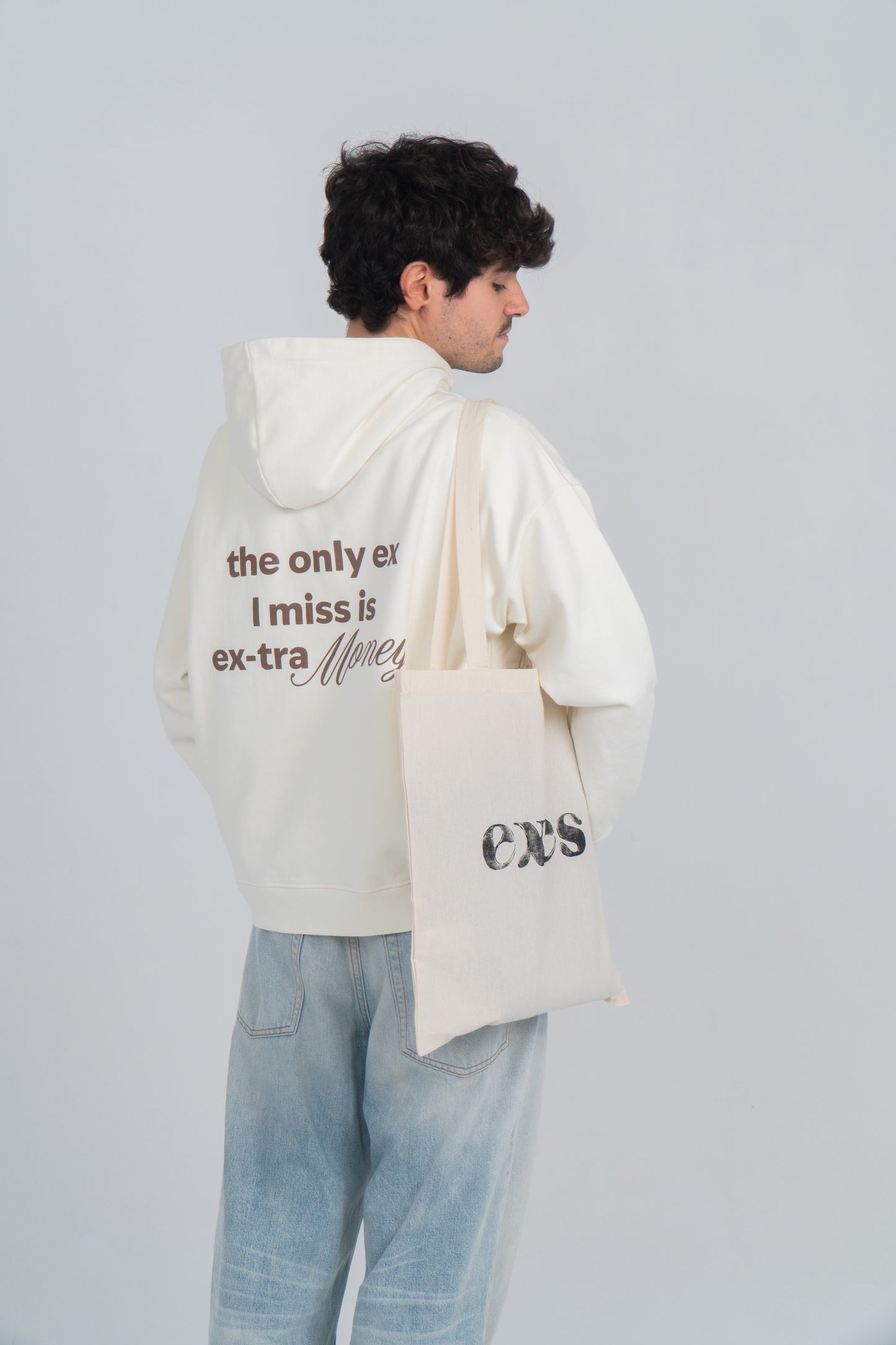 Chico está de espaldas y lleva hoodie/sudadera beige con diseño en la espalda en marrón oscuro que dice "the only ex i miss is ex-tra money". La palabra money está resaltada con otra tipografía. Además el chico lleva colgada la totebag de algodón natural (beige) con el logo de exes en el medio de color negro en serigrafía.