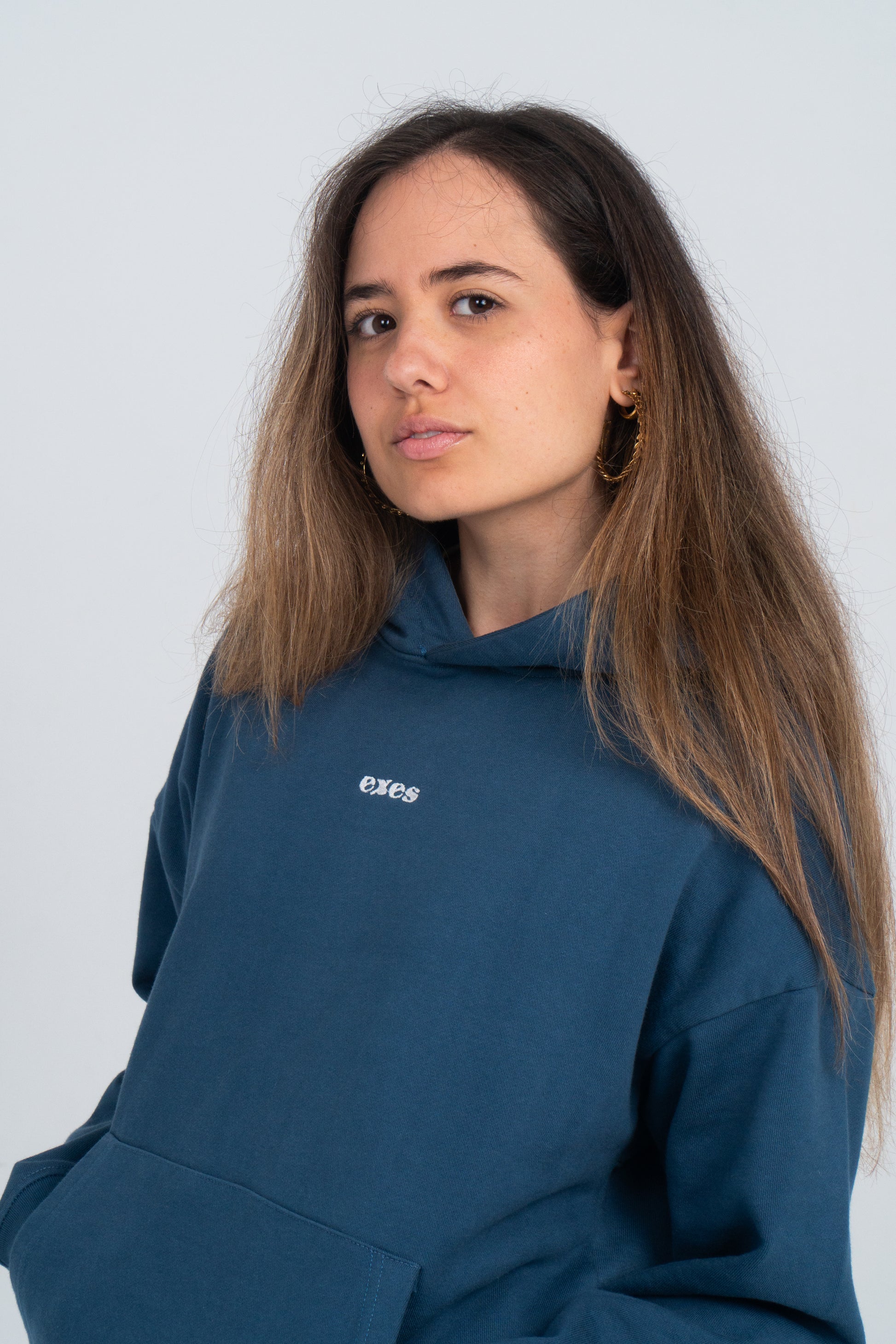 Chica está mirando a cámara. Lleva la sudadera azul del diseño I love You. Tiene el bordado del logo de exes en blanco en el centro del pecho. 